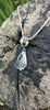 Small triangle-ish mid green seaglass pendant with tendril detail on smooth slim snake chain draped on a mossy woodland log.