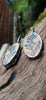 Black silver large drop earrings set with rough sandy coloured stone with black seaweed like inclusions. They lie on a mossy log.