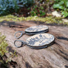 Black silver large drop earrings set with rough sandy coloured stone with black seaweed like inclusions. They lie on a mossy log.