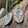 Black silver large drop earrings set with rough sandy coloured stone with black seaweed like inclusions. They lie on a mossy log.