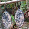 Black silver large drop earrings set with rough sandy coloured stone with black seaweed like inclusions, hang from a twig in the forest.