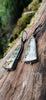Black silver large drop earrings set with an orange green and brown stone, lie in a mossy log in the sunshine. A tine black tendril runs around the outside and a yellow gold dot at the bottom.