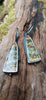 Black silver large drop earrings set with an orange green and brown stone, lie in a mossy log in the sunshine. A tine black tendril runs around the outside and a yellow gold dot at the bottom.