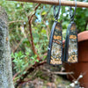 Black silver large drop earrings set with an orange green and brown stone, hang from a twig in the garden. A tine black tendril runs around the outside and a yellow gold dot at the bottom.