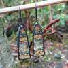 Black silver large drop earrings set with an orange green and brown stone, hang from a twig in the garden. A tine black tendril runs around the outside and a yellow gold dot at the bottom.