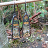 Black silver large drop earrings set with an orange green and brown stone, hang from a twig in the garden. A tine black tendril runs around the outside and a yellow gold dot at the bottom.