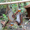 Mismatched dangle earrings with agate slices set with prongs and bezel set amber, hanging from a twig in the garden.