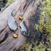 Mismatched dangle earrings with agate slices set with prongs and bezel set amber, lying on a mossy log in the sunshine.