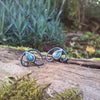Bud shaped with tendril tails curling around, oxidised silver stud earrings with a dreamy elfin quality sits on a mossy log, the labradorite stones glinting blue in the sunlight.