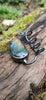 Bud shaped labradorite pendant with a tendril  curling around. on its own with no chain sits on a mossy log, the labradorite stone glinting blue and orange in the sunlight.