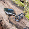 Large Bud shaped Labradorite and oxidised sterling silver pendant sitting on a mossy log, the labradorite stone bud grows out of a tangle of tendrils glinting blue in the sunlight. pendant in it's own no chain.