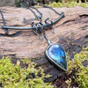 Large Bud shaped Labradorite and oxidised sterling silver pendant sitting on a mossy log, the labradorite stone bud grows out of a tangle of tendrils winding their way around a bark textured bar and chain and glints blue in the sunlight.