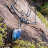 Large Bud shaped Labradorite and oxidised sterling silver pendant sitting on a mossy log, the labradorite stone bud grows out of a tangle of tendrils winding their way around a bark textured bar and chain and glints blue in the sunlight.