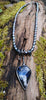 Large Bud shaped Labradorite and oxidised sterling silver pendant sitting on a mossy log, the labradorite stone bud grows out of a tangle of tendrils winding their way around a lustrous strand of black freshwater pearls and glints blue in the sunlight.