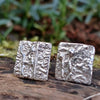  small square melt textured silver studs sitting on a mossy log in the sunshine.