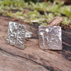 small square melt textured silver studs sitting on a mossy log in the sunshine.