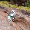 Silver ring with a textured circular top and a small green stone, sitting on a mossy log.