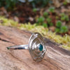 Silver ring with a textured circular top and a small green stone, sitting on a mossy log