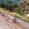 Silver ring with a textured circular top and a small green stone, sitting on a mossy log