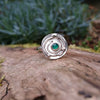 Silver ring with a textured circular top and a small green stone, sitting on a mossy log