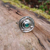 Silver ring with a textured circular top and a small green stone, sitting on a mossy log
