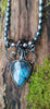 Black oxidised sterling tangle of tendrils wrap around a string of oval shaped black freshwater pearls, terminating in a large bud shaped labradorite with a strong blue flash. The necklace sits on a mossy log in the sunshine.