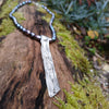 Long sterling silver melt textured pendant on black freshwater pearl beads. the necklace lies on a mossy woodland log.