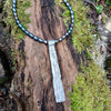 Long sterling silver melt textured pendant on black freshwater pearl beads. the necklace lies on a mossy woodland log.