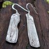 Sterling silver melt textured long drop earrings sitting on a mossy log.