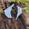 Odd irregular curved triangle shaped melt textured silver studs sitting on a mossy log in the sunshine.