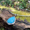 Blue stone pendant with silver tendrils on a silver bar and chain sits on a mossy log