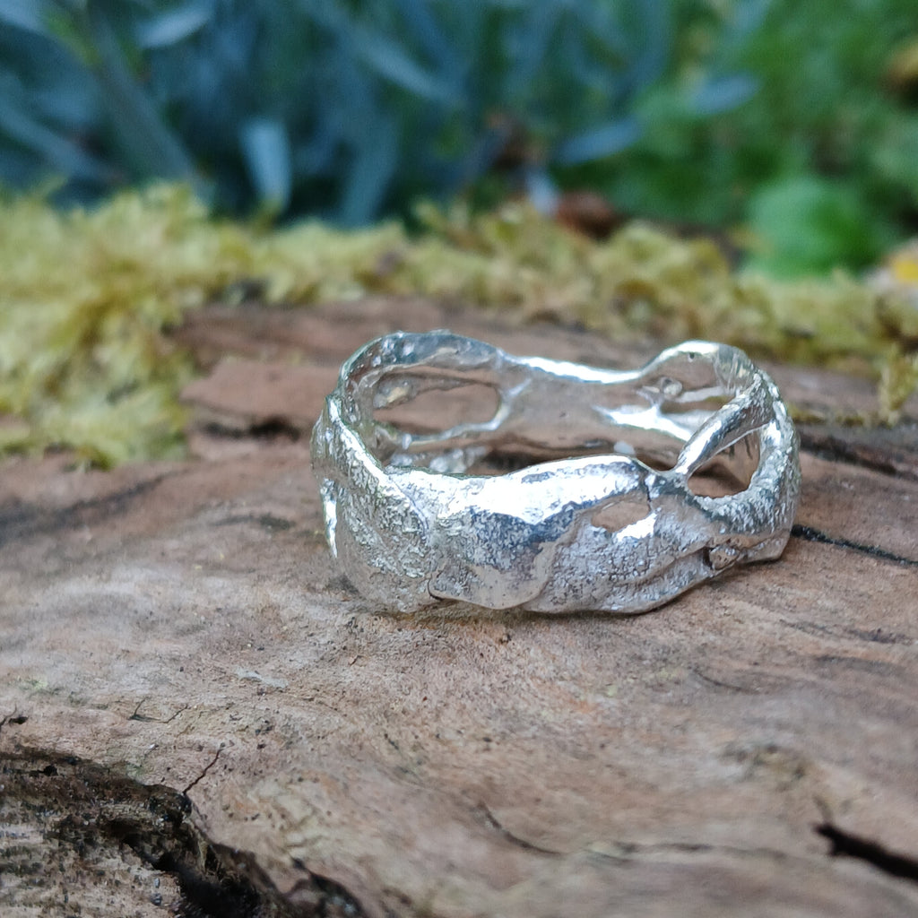 A wibbly wobbly  holey molten textured silver ring sits on a mossy log with leaves in the backround.