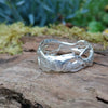  A wibbly wobbly  holey molten textured silver ring sits on a mossy log with leaves in the backround.