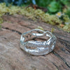 A wibbly wobbly  holey molten textured silver ring sits on a mossy log with leaves in the backround.
