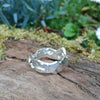 A second wibbly wobbly  holey molten textured silver ring sits on a mossy log with leaves in the backround.