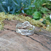A second wibbly wobbly  holey molten textured silver ring sits on a mossy log with leaves in the backround.