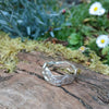 A second wibbly wobbly  holey molten textured silver ring sits on a mossy log with leaves in the backround.