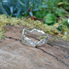 A second wibbly wobbly  holey molten textured silver ring sits on a mossy log with leaves in the backround.