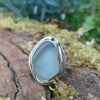 A silver ring with a large, sea glass stone with a muted blue-green hue, shown anngled to see the tendril strand surrounding the stone. It is sitting on a mossy log with blurred green foliage and white daisys in the background.