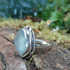 A silver ring with a large, sea glass stone with a muted blue-green hue, shown sitting on a mossy log with blurred green foliage  in the background. Side view showing the tendril wrapping around the stone.