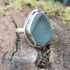 A silver ring with a large, sea glass stone with a muted blue-green hue, shown sitting on a mossy log with blurred green foliage and white daisys in the background.