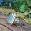 A silver ring with a small sea glass stone with a muted blue-green hue, and a silver tendril wrapping around. shown sitting on a mossy log with blurred green foliage in the background. Side view showing the slim flat band.