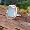 A silver ring with a small sea glass stone with a muted blue-green hue, and a silver tendril wrapping around. shown sitting on a mossy log with blurred green foliage in the background.