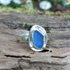 A silver ring with a small blue freeform sea glass stone, and a silver tendril wrapping around. shown sitting on a mossy log with blurred daisys in the background
