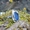 A silver ring with a small blue freeform sea glass stone, and a silver tendril wrapping around. shown sitting on a mossy log with blurred woodland in the background. Side view showing the slim flat band.
