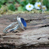 A silver ring with a small blue freeform sea glass stone, and a silver tendril wrapping around. shown sitting on a mossy log with blurred daisys in the background. Side view showing the slim flat band.
