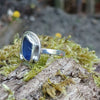 A silver ring with a small blue freeform sea glass stone, and a silver tendril wrapping around. shown sitting on a mossy log with blurred woodland in the background. Side view showing the slim flat band.