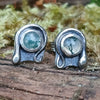 Oxidised finish sterling silver and moss agate stud earrings with tiny tendril wrapping around the stone, sitting on a mossy log in the sunshine.