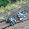 Oxidised finish sterling silver and moss agate stud earrings with tiny tendril wrapping around the stone, sitting on a mossy log in the sunshine.