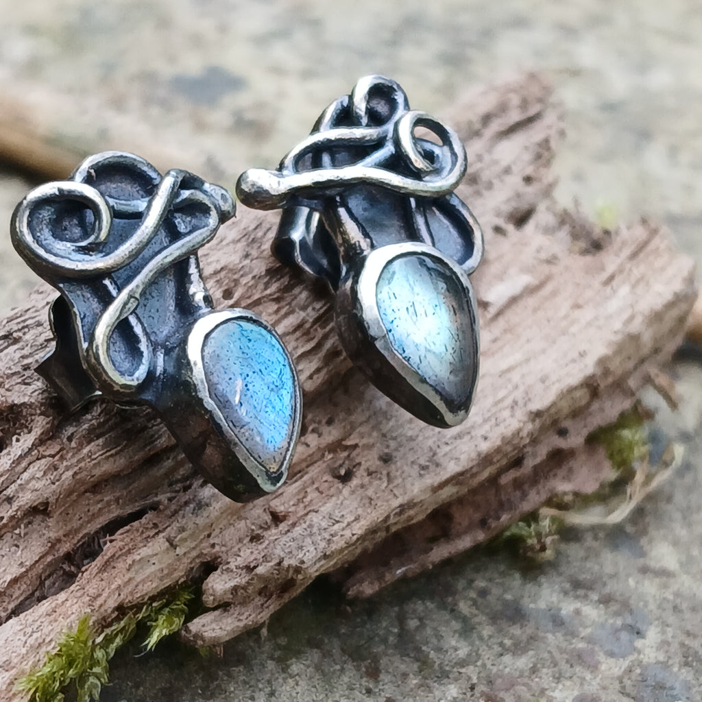 Bud shaped labradorite stones with tangled tendril tails. oxidised silver stud earrings with a dreamy elfin quality sits on a weathered piece of wood, the labradorite stones glinting blue in the sunlight. Shown with the buds facing down and the tendrils up.